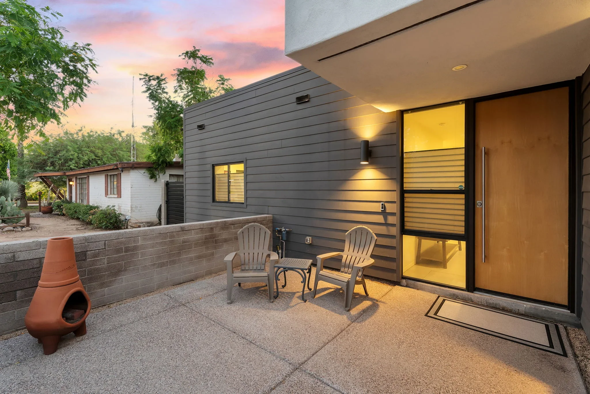 Modern home addition with dark horizontal siding, large windows, wood door, and concrete patio at dusk