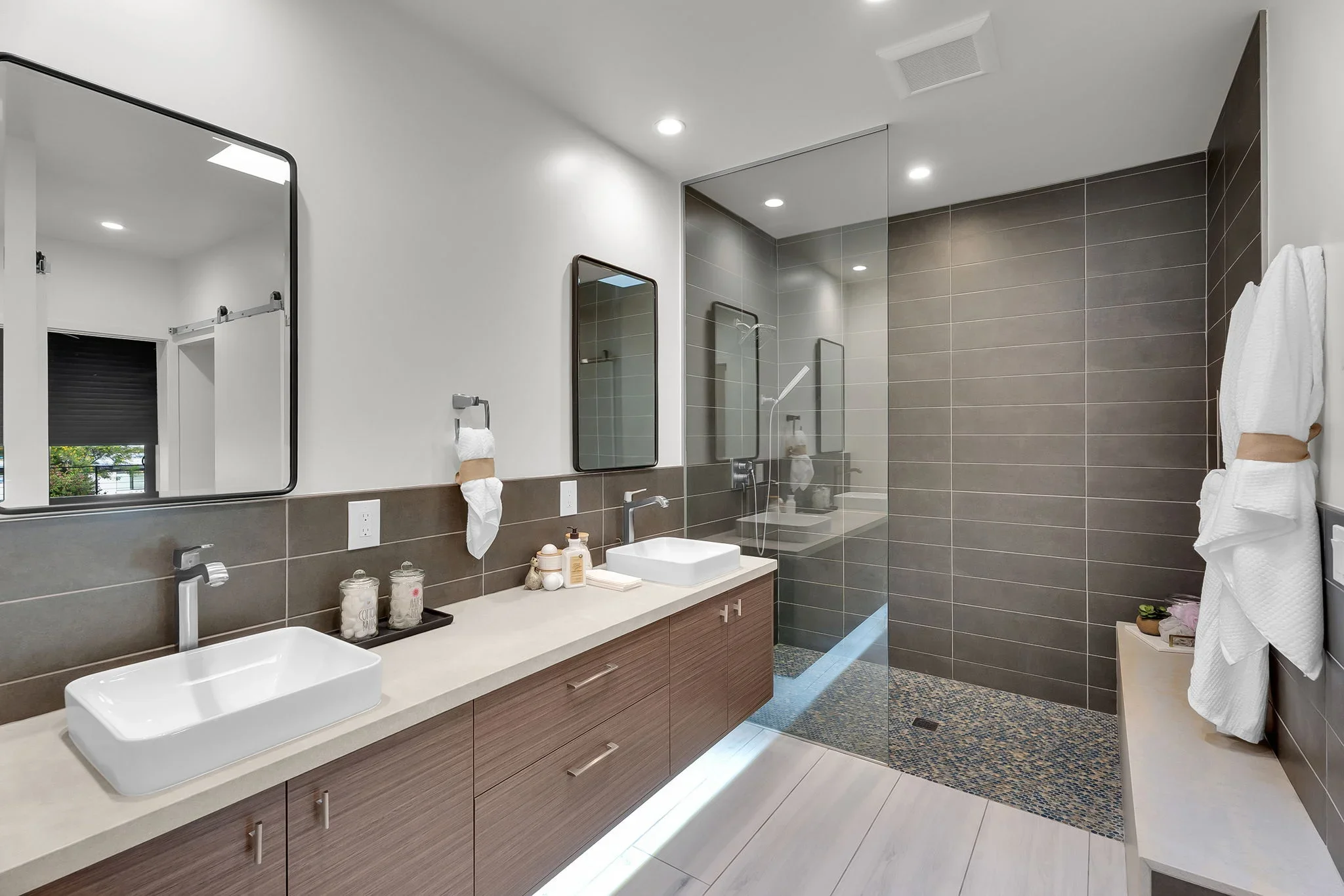 Modern bathroom with wood vanity, dual sinks, gray tile walls, and frameless glass shower enclosure